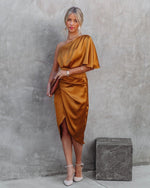 Woman wearing a gold one-shoulder dress against a gray concrete wall.