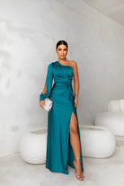Woman in a teal one-shoulder dress with a slit, standing against a white wall.