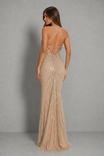 A woman modeling a floor-length champagne beige sequined gown from the back, featuring thin strappy back details and a fitted silhouette, set against a neutral gray studio background.
