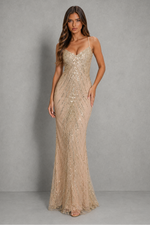A woman modeling a floor-length champagne beige sequined gown with spaghetti straps and a sweetheart neckline, standing against a textured gray studio backdrop.