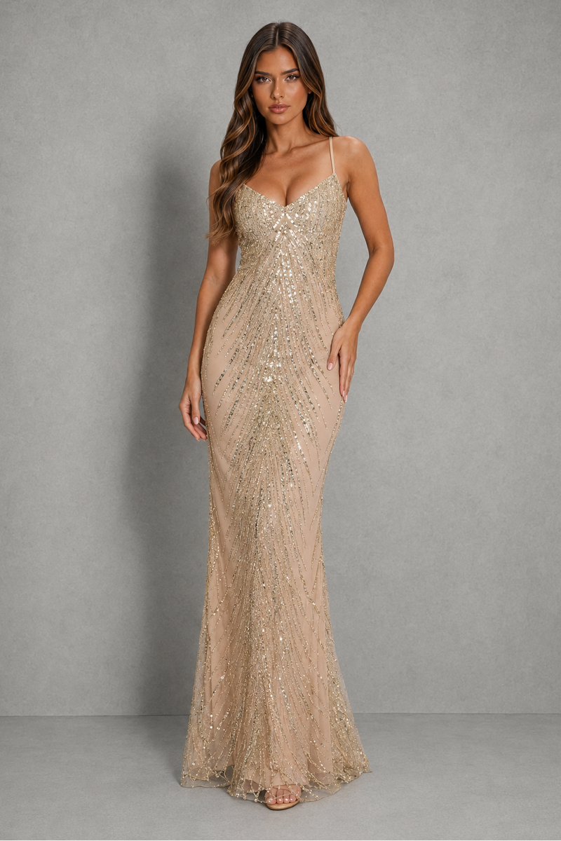 A woman modeling a floor-length champagne beige sequined gown with spaghetti straps and a sweetheart neckline, standing against a textured gray studio backdrop.