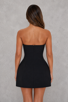 A woman viewed from the back wearing a strapless black mini dress with a structured corset-style waist and a straight skirt, standing against a textured gray wall.
