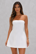 A woman modeling a strapless white mini dress with a structured, corset-style bodice and a flared skirt, set against a textured gray studio background.