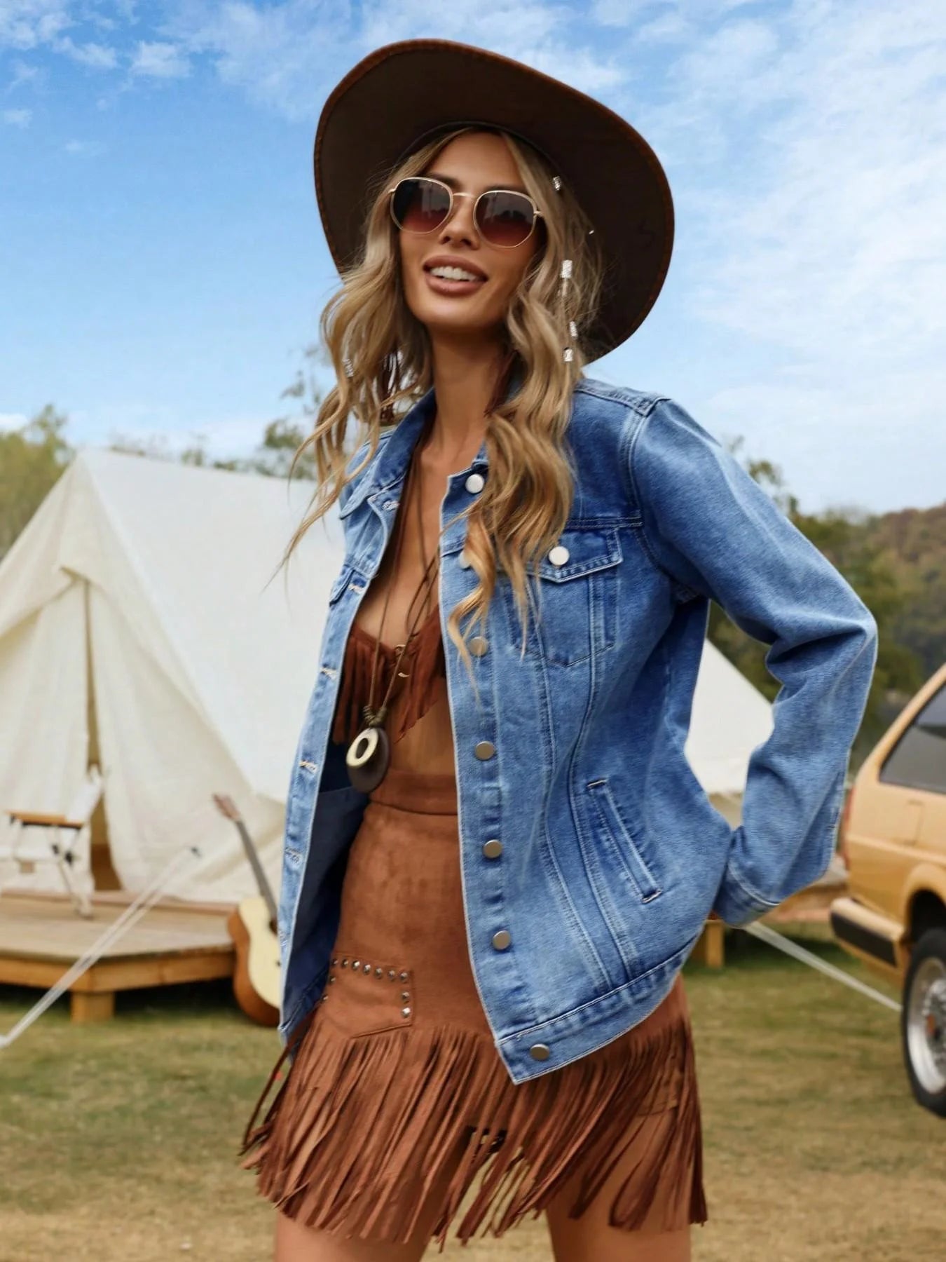 Woman in a denim jacket and brown fringed dress standing outdoors with tents and a car in the background.