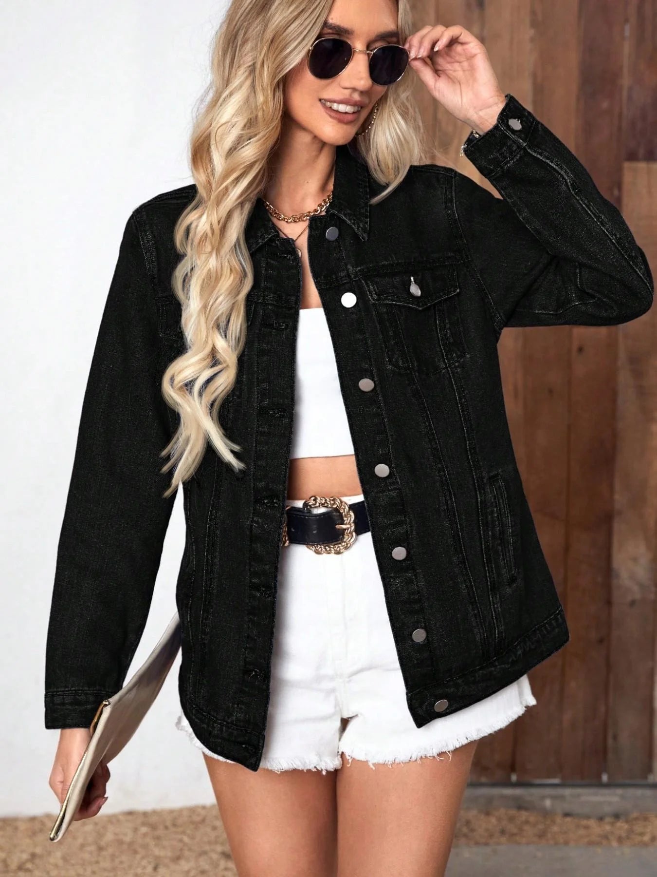 Woman wearing a black denim jacket over a white outfit, holding sunglasses.