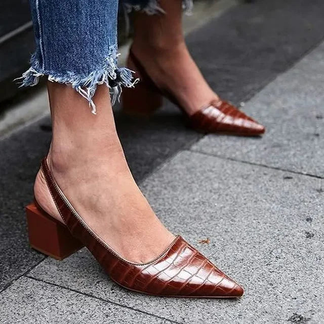 Brown pointed-toe shoes worn with blue jeans on a pavement.
