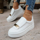 White slip-on sneakers with gold accents worn by a person on a concrete floor.