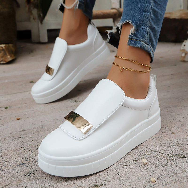 White slip-on sneakers with gold accents worn by a person on a concrete floor.