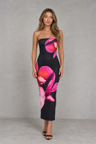 A woman modeling a strapless midi dress with a black base and large pink floral print, standing against a textured gray studio backdrop.