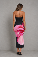 A woman viewed from the back wearing a strapless black midi dress with a large pink flower print on the lower back and hips, paired with black heels, against a gray studio backdrop.