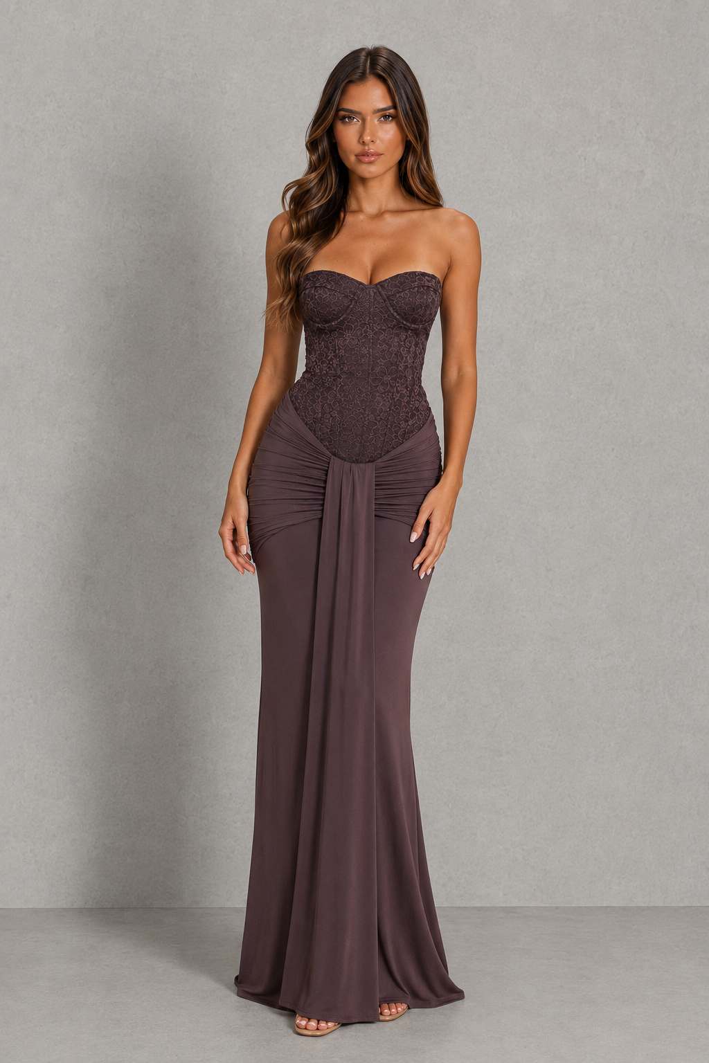 A woman modeling a strapless floor-length gown in a muted plum-brown shade against a neutral gray studio backdrop.