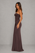 A woman modeling a strapless floor-length gown in a dark chocolate brown color, standing against a neutral gray studio backdrop.