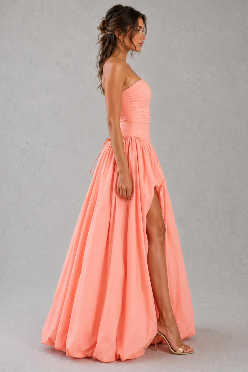 Woman wearing a coral pink strapless gown with a slit on a gray background