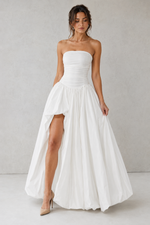 Woman wearing a strapless white wedding dress with a high slit on a plain background