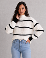 Woman wearing a white sweater with black stripes and blue jeans against a gray background
