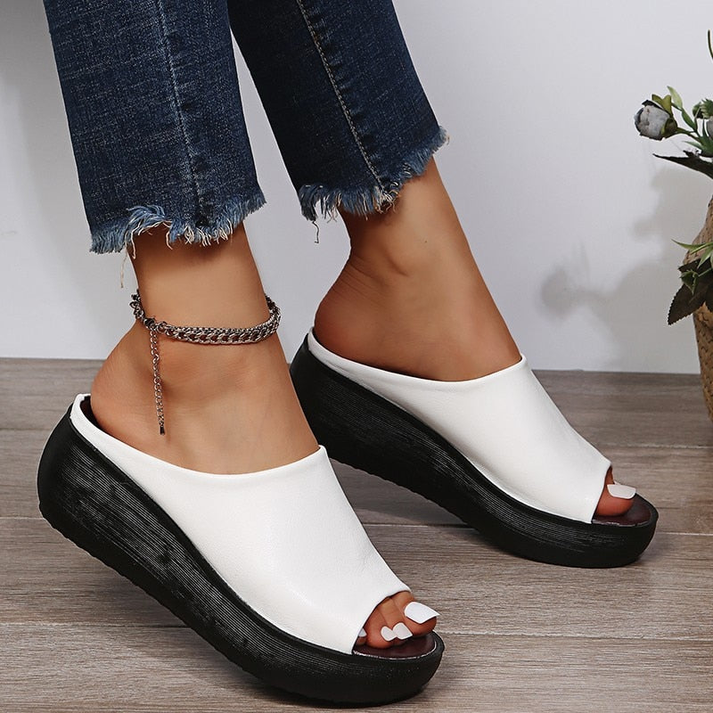 White open-toe sandals with black soles worn by a person on a wooden floor.