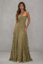 A woman modeling a floor-length olive green strapless gown with a fitted bodice and sheer floral lace side panels, standing against a textured gray studio backdrop.