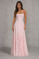 A woman modeling a floor-length light pink gown with thin straps, a fitted ruched bodice, and a flared skirt, standing against a textured gray studio backdrop.