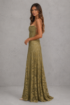 A woman modeling a strapless olive green lace maxi gown with a fitted bodice and flowing skirt, standing against a textured gray studio backdrop.