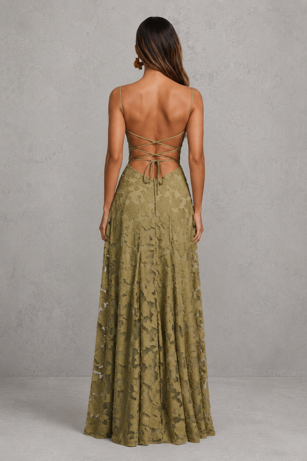 A woman shown from the back wearing a sleeveless olive green lace maxi dress with a strappy open back, standing against a textured gray studio wall.