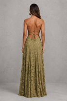 A woman shown from the back wearing a sleeveless olive green lace maxi dress with a strappy open back, standing against a textured gray studio wall.