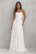 A woman modeling a floor-length white lace dress with thin straps and a fitted bodice, standing against a textured light gray studio backdrop.