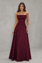 A woman modeling a floor-length burgundy lace gown with thin spaghetti straps, a structured sweetheart neckline, and a full A-line skirt, set against a textured gray studio backdrop.