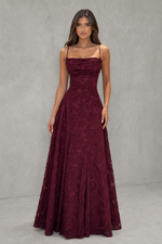 A woman modeling a floor-length burgundy lace gown with thin spaghetti straps, a structured sweetheart neckline, and a full A-line skirt, set against a textured gray studio backdrop.