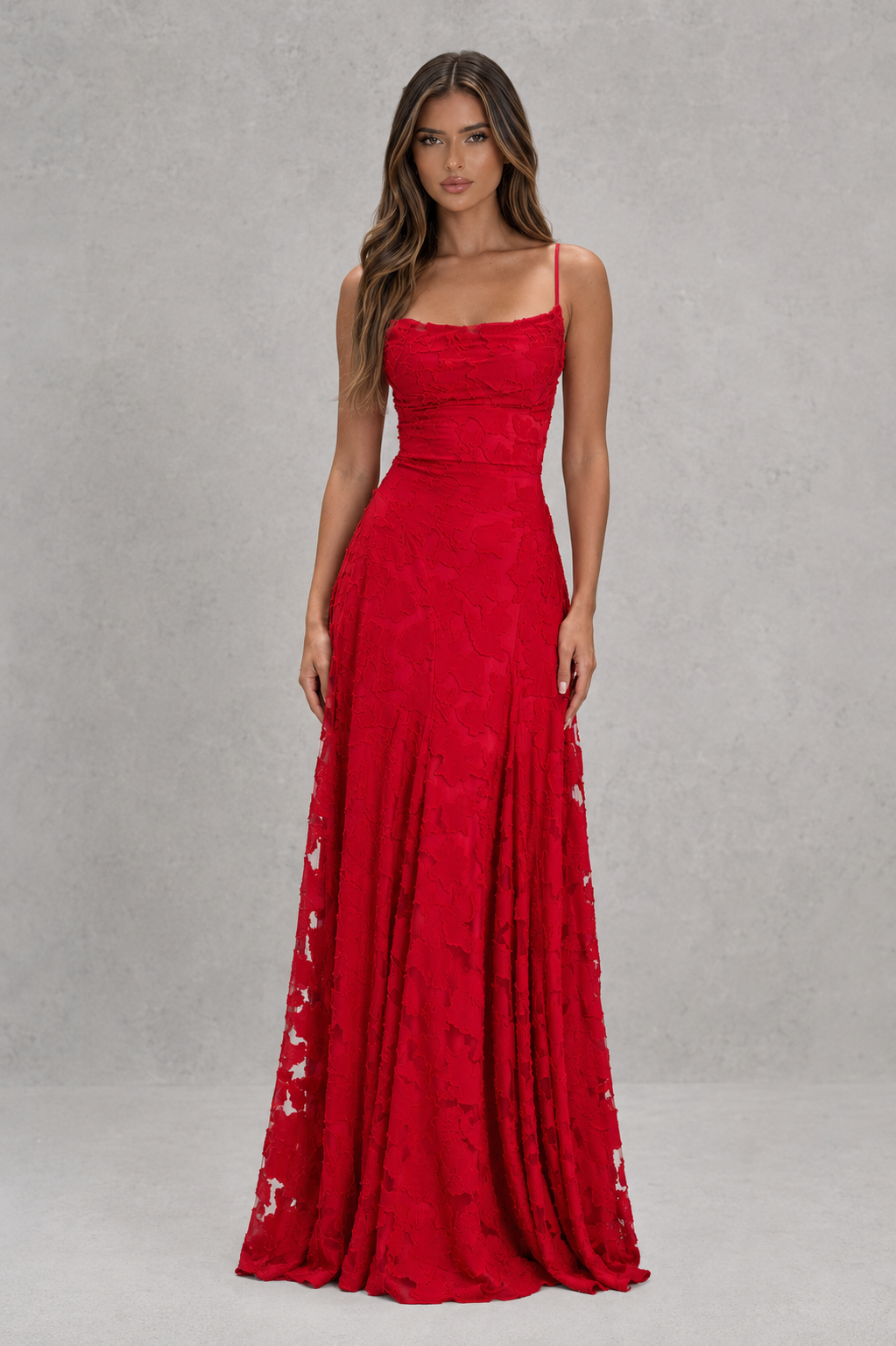 A woman modeling a floor-length red evening gown with thin straps, a fitted waist, and lace-up side details, photographed against a textured gray studio backdrop.