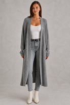 Woman wearing a long plaid cardigan, white top, jeans, and white boots on a gray background