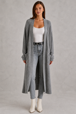 Woman wearing a long plaid cardigan, white top, jeans, and white boots on a gray background