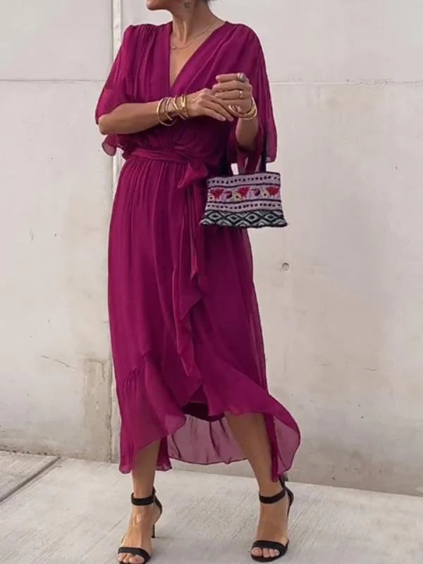Woman wearing a magenta dress with a patterned handbag against a white wall.