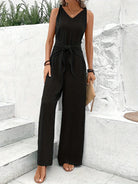 Woman wearing a black jumpsuit standing on stone steps with a white wall and greenery in the background.