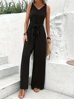 Woman wearing a black jumpsuit standing on stone steps with a white wall and greenery in the background.