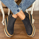 Black slip-on shoes with beige soles worn by a person sitting on a wooden floor.