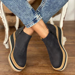 Black slip-on shoes with beige soles worn by a person sitting on a wooden floor.