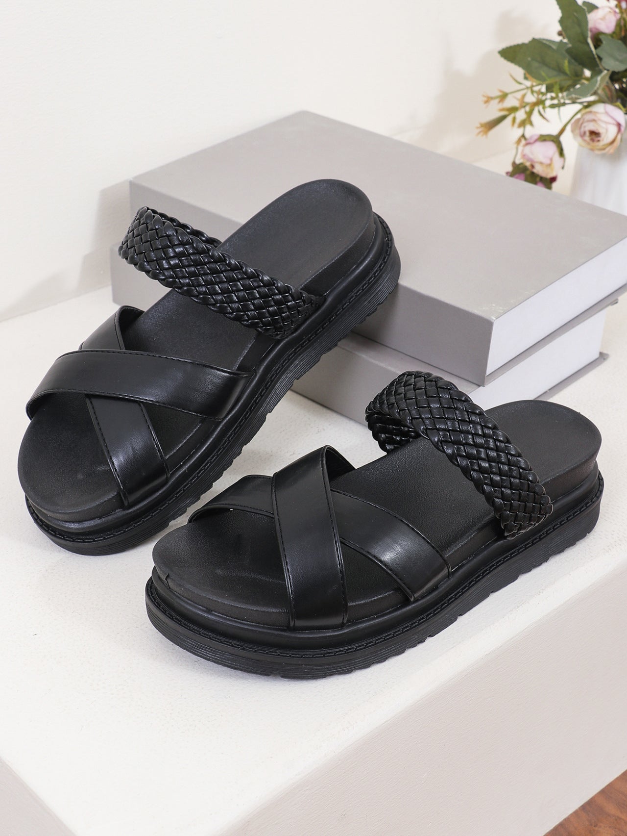 Black sandals with woven straps on a white surface with a gray box and flowers in the background.