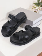 Black sandals with woven straps on a white surface with a gray box and flowers in the background.