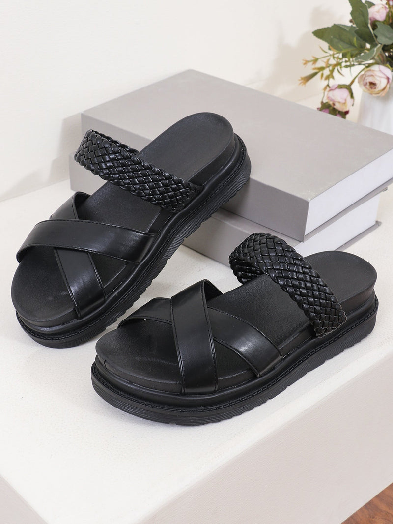 Black sandals with woven straps on a white surface with a gray box and flowers in the background.
