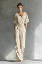 Woman wearing a beige jumpsuit standing against a textured gray wall.