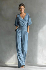 Woman wearing a blue jumpsuit standing against a gray wall.
