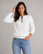 Woman wearing a white cable knit sweater and blue jeans against a gray background